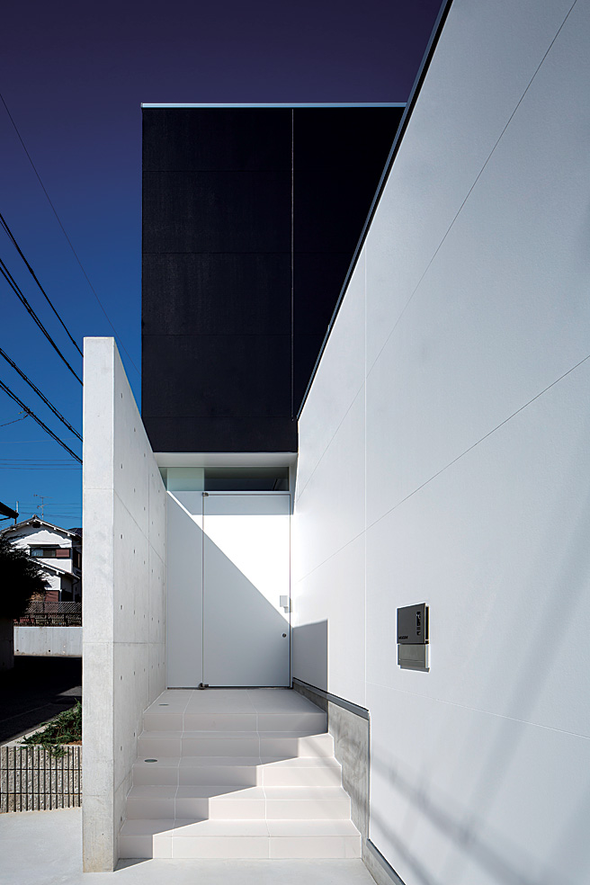 ส่วนของบ้านยกพื้นให้สูงขึ้น เป็นบันไดขึ้นไปหลายขั้น ใช้สีขาวเป็นหลักทำให้ดูสะอาดสะอ้าน ขวา / ลองมองเข้าไปจากทางเข้า จะเห็นกำแพงคอนกรีตและช่องหน้าต่างแบบ slit นอกจากจะเป็นดีไซน์ที่สวยแล้ว ยังเป็นช่องให้แสงแดดส่องเข้ามาได้ ซึ่งเป็นสิ่งสำคัญอีกด้วย